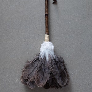 May include: A brown feather duster with a long wooden handle and a leather strap. The duster has a white tip and is made of natural feathers.