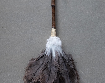 May include: A brown feather duster with a long wooden handle and a leather strap. The duster has a white tip and is made of natural feathers.