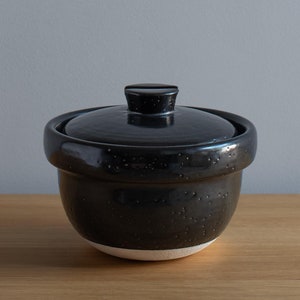 May include: A black ceramic rice cooker with a lid. The cooker has a white rim around the bottom.