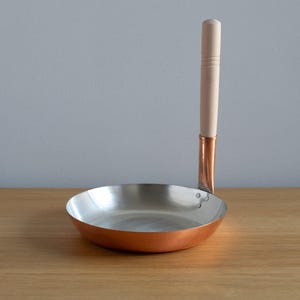 Puede incluir: Una pequeña sartén con fondo de cobre, interior de acero inoxidable y un mango largo de madera de color claro. La sartén es redonda y tiene una superficie pulida y reflectante. El mango está sujeto con un accesorio de cobre. La sartén está diseñada para cocinar.