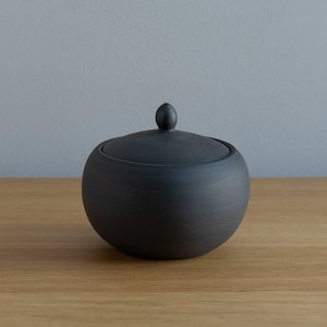 May include: A black ceramic sugar bowl with a lid and a small knob on top. The bowl has a rounded shape and sits on a wooden surface. The background is a solid gray color.