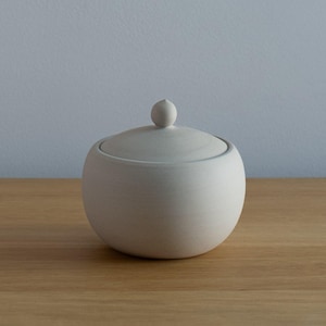 May include: A small, off-white ceramic sugar bowl with a lid and a small spherical handle. The bowl has a rounded shape and sits on a wooden surface. The background is a solid light blue.