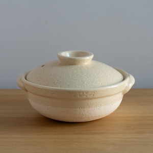 Puede incluir: Una olla japonesa de cerámica beige, llamada donabe, con su tapa a juego. La olla tiene forma redonda, con dos asas y un pequeño pomo en la tapa. La superficie tiene un aspecto texturizado.