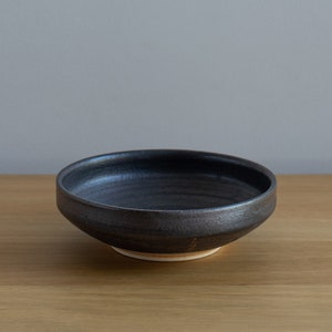 Puede incluir: Un cuenco de cerámica negro con un borde ligeramente elevado y un acabado liso y brillante. El cuenco está sobre una superficie de madera.