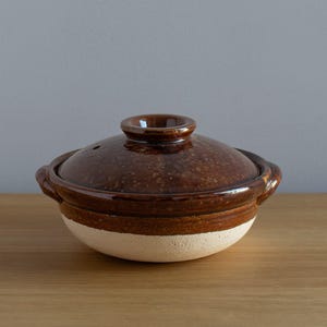 Könnte beinhalten: Ein brauner Keramikkochtopf mit Deckel. Der Topf hat ein weißes Band um den Boden und zwei kleine Griffe. Der Deckel hat einen kleinen Knopf und ein Entlüftungsloch. Der Topf steht auf einer Holzoberfläche.