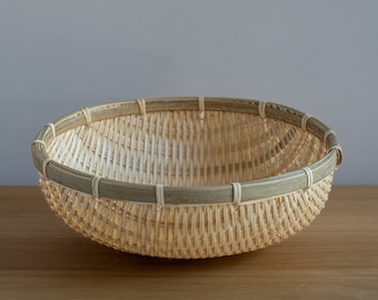 May include: A woven bamboo basket with a natural brown color and a light green rim. The basket is empty and sitting on a wooden surface.