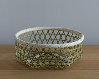 May include: A woven basket made of natural materials, with a light green color and a white rim. The basket has a diamond pattern and is sitting on a wooden surface.