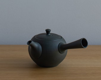 May include: A black ceramic teapot with a long spout and a round body. The teapot is sitting on a wooden surface.