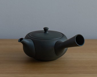 May include: A matte black ceramic teapot with a rounded body, a lid with a small knob, and a long, cylindrical spout. The teapot is designed for tea brewing and sits on a wooden surface.