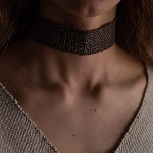 May include: A dark brown woven choker necklace. The choker is made of a textured material and sits around the neck. The neckline of a light beige jumper is visible.