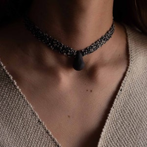 May include: A black choker necklace with a teardrop-shaped pendant. The necklace is made of braided cord and has a simple, minimalist design.