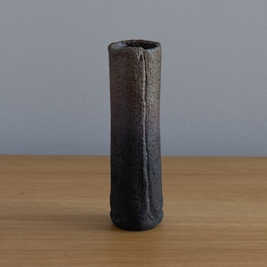 May include: A tall, cylindrical ceramic vase with a textured surface. The vase transitions from a dark, charcoal colour at the base to a lighter, brownish-grey near the top. The vase is approximately 20 cm tall.