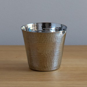 Può includere: Una tazza in metallo argentato martellato con fondo arrotondato e bordo leggermente svasato.