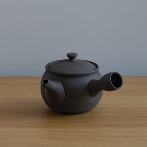 May include: A dark brown ceramic teapot with a rounded body, a lid with a small knob, and a curved spout. The teapot has a textured surface and is designed for brewing tea.