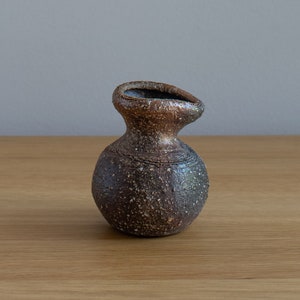 May include: A small, brown, speckled ceramic vase with a rounded body and a slightly uneven opening. The vase has a rustic, handmade look.