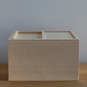 Puede incluir: Una caja de almacenamiento de madera clara con una tapa con bisagras y un divisor en el interior. La caja está cerrada y se encuentra sobre una superficie de madera.