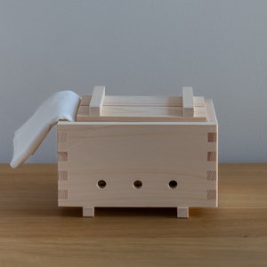 May include: A wooden tofu press with a white cloth draped over the top. The press has a rectangular shape with a lid and a series of holes in the bottom.