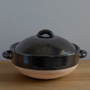 Könnte beinhalten: Ein schwarzer Keramikkochtopf mit passendem Deckel. Der Topf hat einen breiten, abgerundeten Boden mit einem beigefarbenen Band am Boden. Der Deckel hat einen Schlaufengriff und ein kleines Entlüftungsloch. Der Topf hat zwei seitliche Griffe.