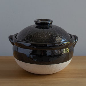 Könnte beinhalten: Ein schwarzer Keramikkochtopf mit passendem Deckel. Der Topf hat eine strukturierte Oberfläche auf dem Deckel und eine glänzende Oberfläche am Rand und an den Griffen. Die untere Hälfte des Topfes ist hellbeige.