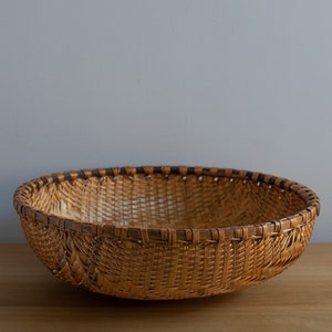 May include: A round, woven basket with a wide, shallow bowl shape. The basket is made of natural-colored reeds with a dark brown rim. The weave is tight and intricate, creating a textured surface. The basket sits on a wooden surface.