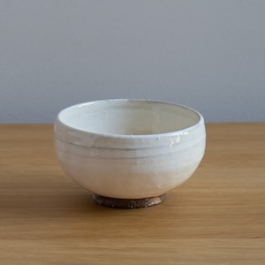 May include: A handmade white ceramic bowl with a slightly textured surface. The bowl has a rounded shape with a wide opening and a dark brown base. The bowl is sitting on a wooden surface.