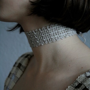 May include: A close-up of a person wearing a light brown woven choker necklace. The necklace is made of a thick, textured material and has a simple, elegant design.