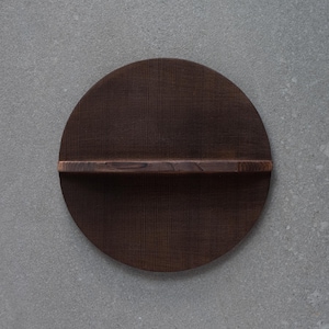Könnte beinhalten: Ein runder, dunkelbrauner Holzregal mit einem horizontalen Regalansatz. Das Regal hat eine geriffelte Textur und ist vor einem grauen Hintergrund angeordnet. Das Regal hat einen Durchmesser von etwa 30 cm.