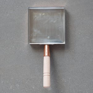 Może przedstawiać: Kwadratowe, metalowe narzędzie kuchenne z drewnianym uchwytem. Metalowa część to płytki, otwarty kwadrat, a uchwyt to jasne drewno z miedzianym metalowym łącznikiem. Narzędzie jest przeznaczone do gotowania.