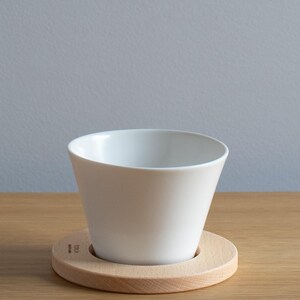 Può includere: Una tazza in ceramica bianca su un sottobicchiere in legno chiaro. La tazza ha una forma svasata, più larga nella parte superiore che nella base. Il sottobicchiere è rotondo con un centro incassato per tenere la tazza. Lo sfondo è un muro grigio.
