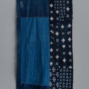Puede incluir: Textil rectangular con diseño de patchwork en tonos azul índigo. El textil presenta varios patrones geométricos y texturas, incluyendo detalles cosidos y paneles de tela contrastantes. La estética general es rústica y artesanal.