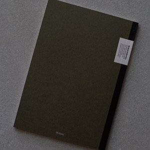 May include: A dark green notebook with a black spine and a white label on the back that says "kleid stationery". The label also includes the text "designed in london, made in italy, 100% recycled paper, 100gsm, 96 pages, 148 x 210mm". The notebook has the word "kleid" printed in white on the front cover.