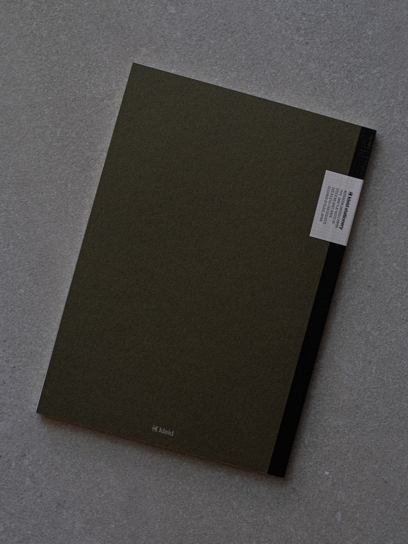 May include: A dark green notebook with a black spine and a white label on the back that says "kleid stationery". The label also includes the text "designed in london, made in italy, 100% recycled paper, 100gsm, 96 pages, 148 x 210mm". The notebook has the word "kleid" printed in white on the front cover.