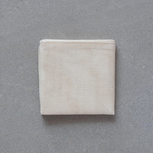 May include: A folded, square, off-white cloth napkin. The napkin is made of a woven material, and the edges are neatly hemmed. The background is a solid, light grey surface.