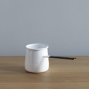 Könnte beinhalten: Eine weiße Emaille-Kaffeekanne mit schwarzem Griff. Die Kanne steht auf einer Holzoberfläche.