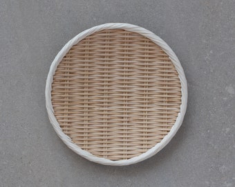 May include: A round, woven bamboo tray with a white rim. The tray is on a gray concrete background.