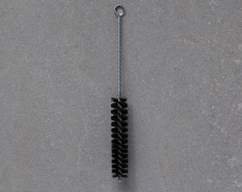 May include: A bottle brush with a black bristled head and a silver metal handle. The handle has a loop at the top. The brush is designed for cleaning bottles and other narrow-necked containers.