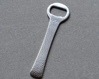 May include: A silver bottle opener with a textured handle and a loop at the top. The opener is made of metal and has a hammered design on the handle. The design is simple and modern.