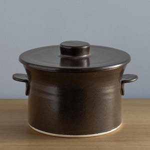 Könnte beinhalten: Ein dunkelbrauner Keramikkochtopf mit Deckel und zwei seitlichen Griffen. Der Topf hat eine abgerundete Form mit einem weißen Rand am Boden. Der Deckel hat einen kleinen, runden Griff oben. Der Topf steht auf einer Holzoberfläche.