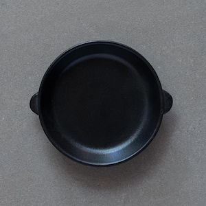 Pode incluir: Uma frigideira de ferro fundido preta com duas alças pequenas. A frigideira está vazia e está sobre uma superfície cinza.