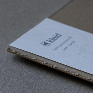 May include: A close-up of a brown cardboard box with a white label. The label has the word "Kleid" printed on it, along with the text "2mm grid of dots, colour: Camel".