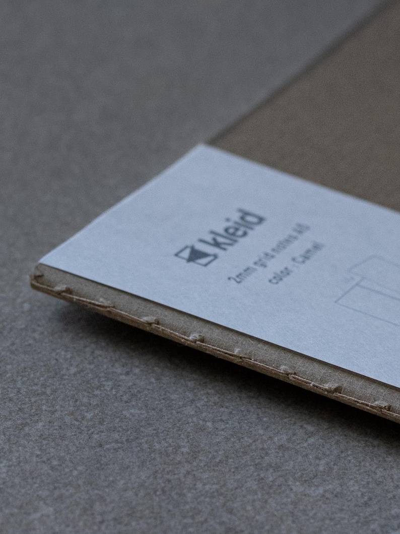 May include: A close-up of a brown cardboard box with a white label. The label has the word "Kleid" printed on it, along with the text "2mm grid of dots, colour: Camel".