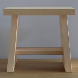 May include: A natural wood stool with a simple design. The stool has a rectangular top and a shelf underneath. The stool is made of light wood and has a natural finish.