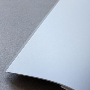May include: A blank, white notebook with a slightly curved spine. The notebook is open and lying flat on a grey surface.