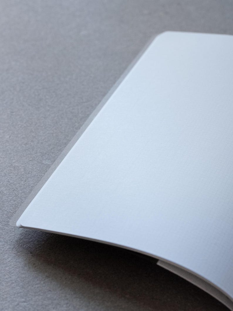 May include: A blank, white notebook with a slightly curved spine. The notebook is open and lying flat on a grey surface.