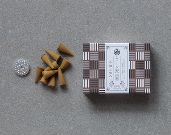 May include: A box of incense cones with a geometric pattern in brown, grey, and white, next to a small metal incense holder and a pile of brown incense cones. The box has Japanese text. The items are on a grey surface.