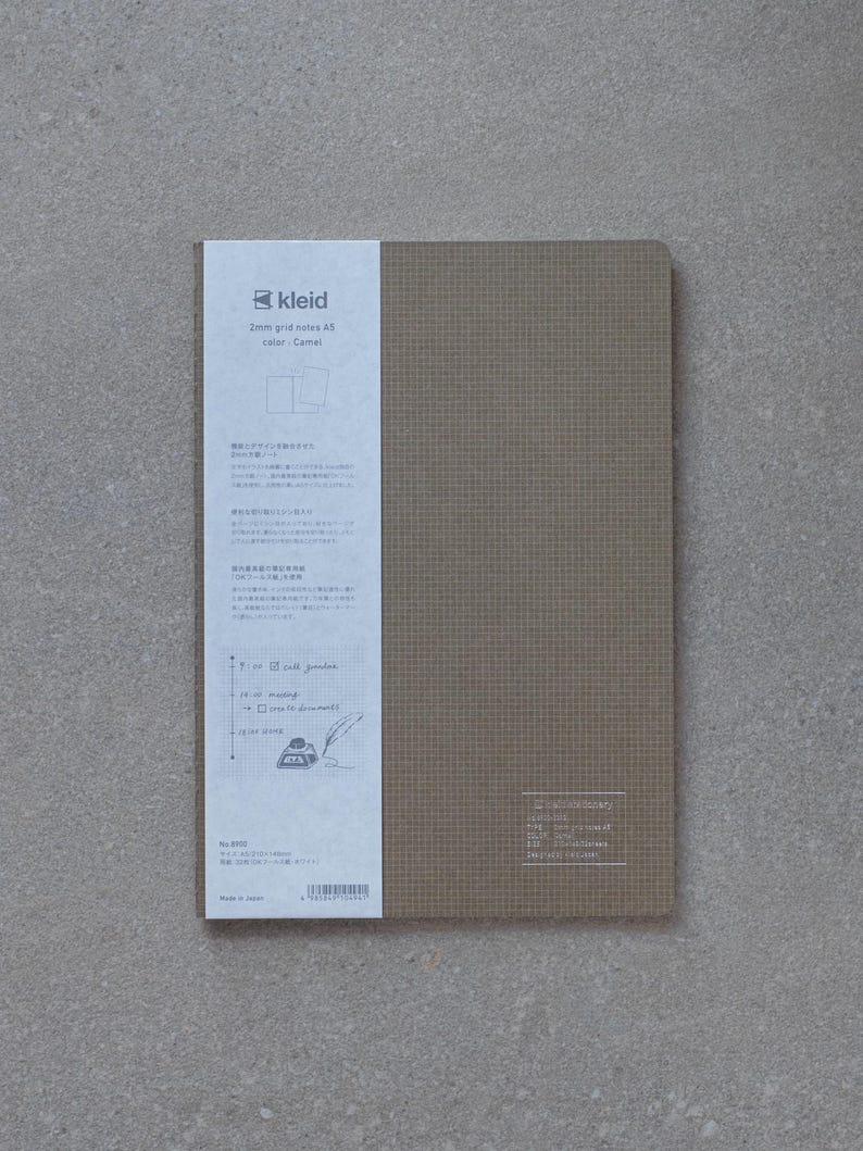 May include: A brown A5 notebook with a 2mm grid. The cover has a subtle texture and the text "kleid 2mm grid notes A5 color: Camel" is printed on the front. The back of the notebook has Japanese text and a small illustration of a quill pen and inkwell.