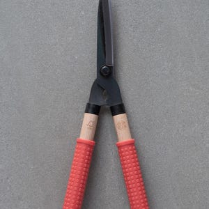 May include: A pair of black garden shears with wooden handles and red rubber grips. The blades are sharp and pointed, designed for precise cutting. The shears are set against a gray background.
