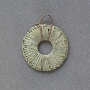 May include: A round, woven, straw coaster in a natural beige colour. The coaster has a hole in the centre and is tied with a brown twine.