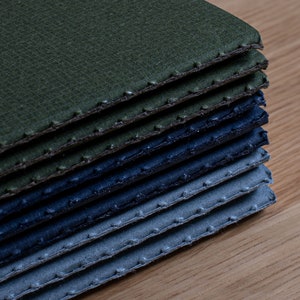 May include: A stack of fabric swatches in shades of green, blue, and grey. The fabric has a textured surface and is stitched around the edges.