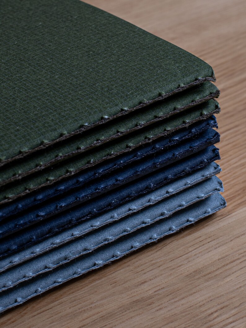 May include: A stack of fabric swatches in shades of green, blue, and grey. The fabric has a textured surface and is stitched around the edges.